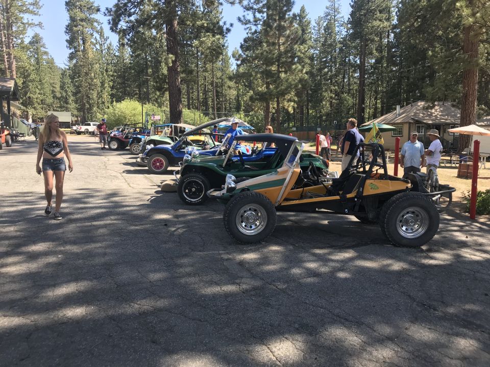 Several dune buggies parked in a lot with trees and buildings in the background. A woman walks in the foreground