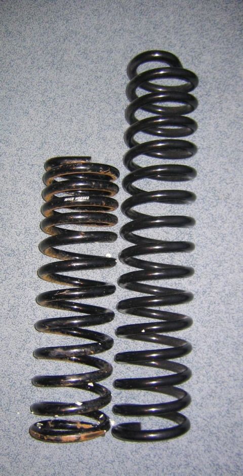 1978 Ford Bronco - Two front coil springs laid side by side on gray carpet for a height comparison: a rusty, paint-chipped used spring on the left with a WILD…
