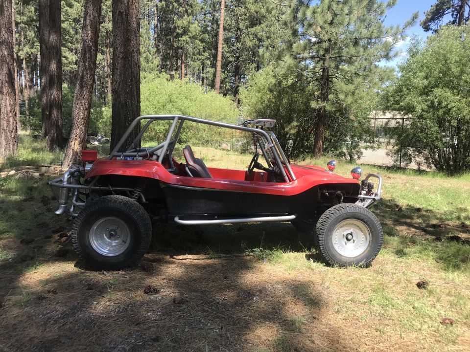 Red and black dune buggy with chrome roll cage parked in a wooded area.