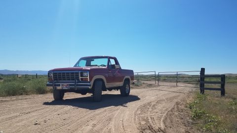 1985 Ford Bronco - A red and tan 1980s pickup truck is parked on a dirt road in front of a metal gate and fence. The truck is facing the camera at a slight an…