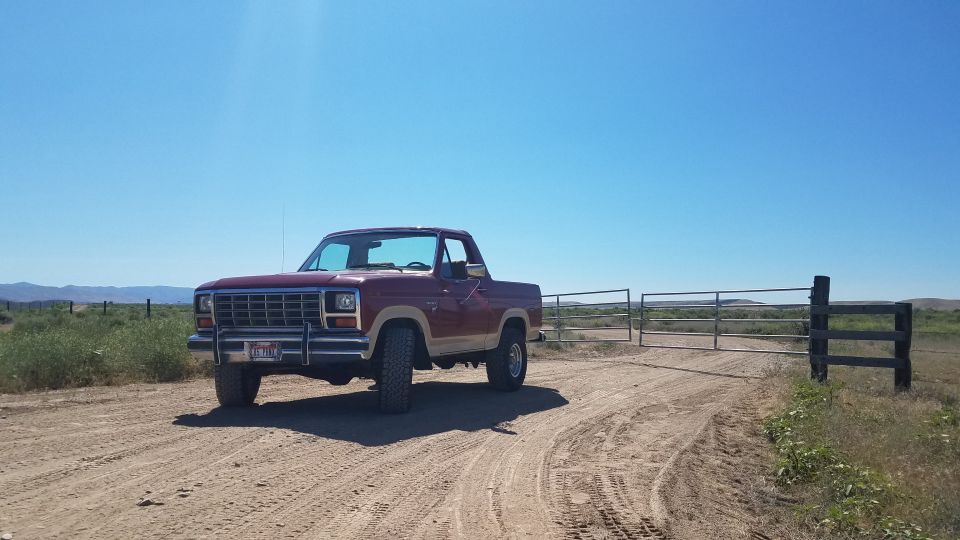 Red and tan 1980s pickup truck parked on a dirt road near a gate.