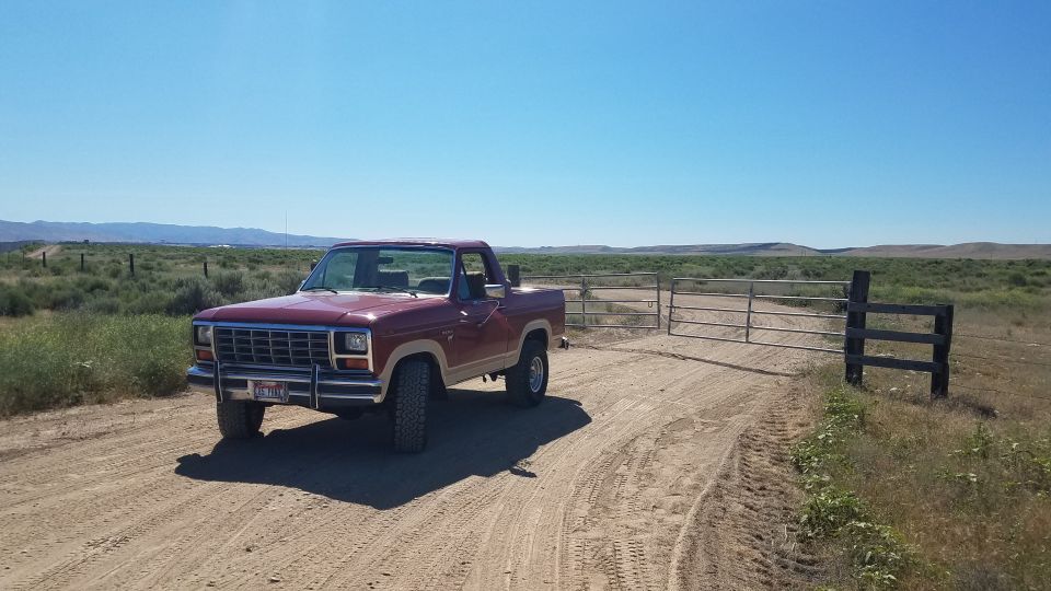 Maroon and tan 1980s Ford pickup truck parked on a dirt road next to a metal gate.