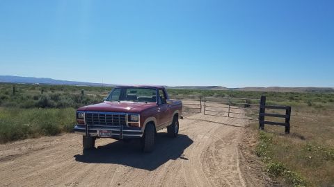 1985 Ford Bronco - A red 1980s pickup truck is parked on a dirt road next to a metal gate in a dry, grassy landscape under a clear blue sky.