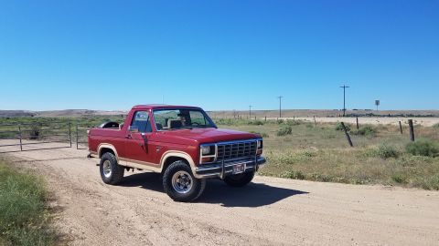 1985 Ford Bronco - A red and tan 1985 Ford Bronco is parked on a dirt road in a dry, rural landscape under a clear blue sky. The vehicle is viewed from a 3/4…
