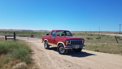 1985 Ford Bronco - A red 1980s Ford Bronco is parked on a dirt road in front of a metal gate on a sunny day. The vehicle is facing slightly left, with a spare…