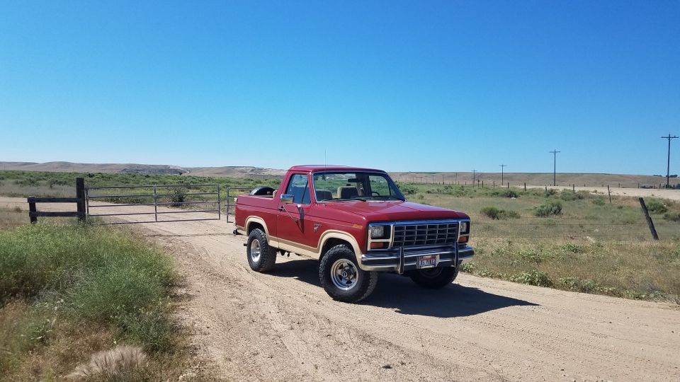 Red 1980s Ford Bronco parked on a dirt road near a gate.