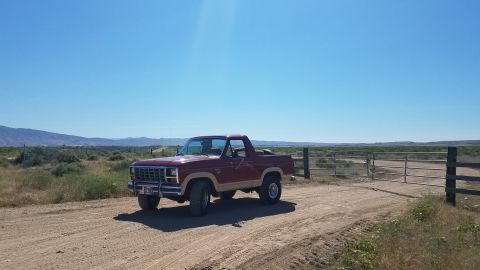 1985 Ford Bronco - A maroon and tan 1980s Ford Bronco is parked on a dirt road in a dry, rural landscape with mountains in the distance.