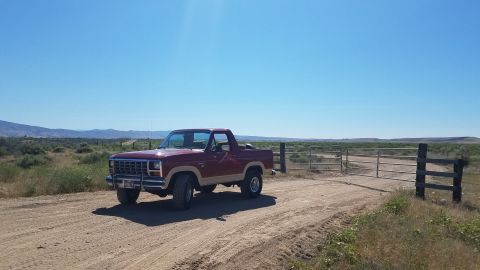 1985 Ford Bronco - A maroon and tan 1985 Ford Bronco is parked on a dirt road next to a metal gate in a rural landscape under a clear blue sky.