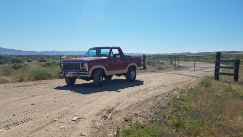 1985 Ford Bronco - A maroon and tan two-door Ford Bronco is parked on a dirt road next to a metal gate in a dry, rural landscape.