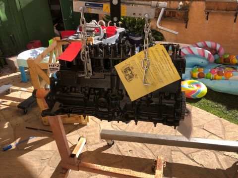 1992 Jeep Wrangler - A black engine block is suspended by chains from an engine hoist. A yellow instruction manual is attached to the engine, and a red tag is a…