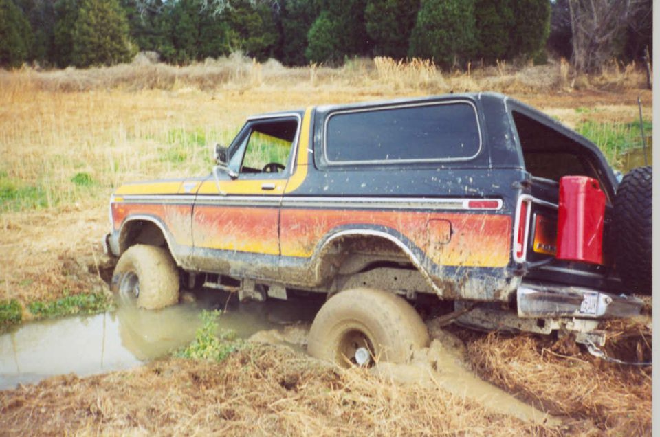 Black Ford Bronco with orange sunburst stripes stuck axle-deep in a muddy field