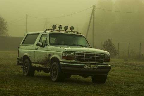 1993 Ford Bronco - A white, muddy Ford Bronco is parked on a grassy field with a foggy, overcast sky in the background. The vehicle has a roof rack with four…