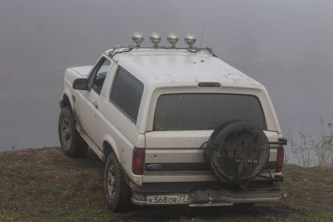 1993 Ford Bronco - The rear-driver side quarter view of a white Ford Bronco is parked on a grassy, muddy incline next to a body of water on a foggy day. The v…