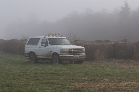 1993 Ford Bronco - A white Ford Bronco is parked in a grassy field next to large hay bales on a foggy day. The vehicle has four auxiliary lights mounted on th…