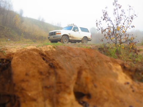 1993 Ford Bronco - A white 1993 Ford Bronco is parked on a dirt path with a muddy hill in the foreground and a misty, wooded hillside in the background. The v…