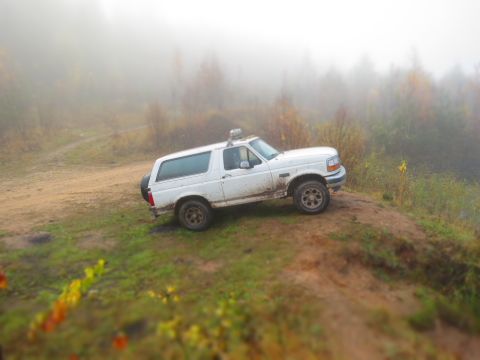 1993 Ford Bronco - A white, muddy Ford Bronco is parked on a dirt hill with trees and fog in the background. The vehicle has a spare tire mounted on the rear.