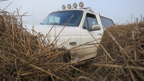 1993 Ford Bronco - A white Ford Bronco is partially obscured by dry reeds and vegetation. The vehicle has four auxiliary lights mounted on its roof rack and a…