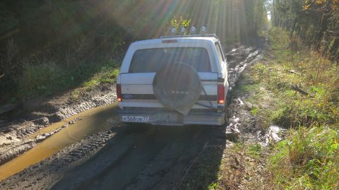 1993 Ford Bronco - The rear of a white Ford Bronco is shown driving on a muddy, rutted dirt road through a forest. The vehicle has a spare tire mounted on the…