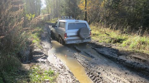 1993 Ford Bronco - A silver Ford Bronco is driving through a muddy, rutted path in a forest. The vehicle is partially submerged in a large puddle of water.