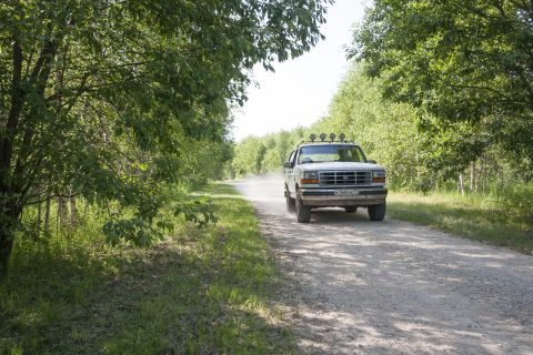1993 Ford Bronco - A white Ford Bronco drives down a dusty dirt road surrounded by trees. The vehicle has four auxiliary lights mounted on the roof rack.