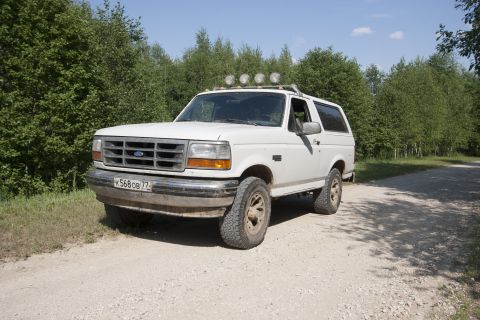 1993 Ford Bronco - A white 1993 Ford Bronco is parked on a dirt road surrounded by trees. The vehicle has four auxiliary lights mounted on the roof rack and a…