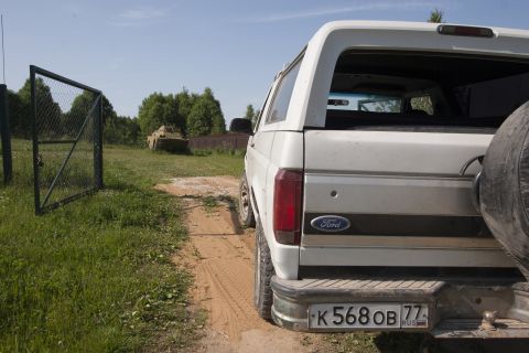 1993 Ford Bronco - The rear of a white Ford Bronco is parked on a dirt road next to a grassy field. A metal gate and a small armored vehicle are visible in th…