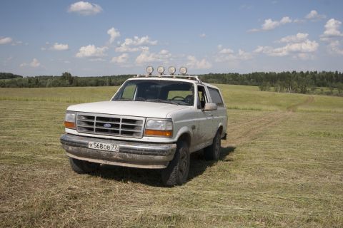 1993 Ford Bronco - A white 1993 Ford Bronco is parked in a mown grassy field under a blue sky with scattered clouds. The vehicle has a roof-mounted light bar…