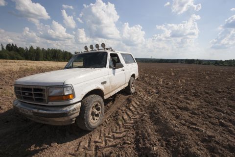 1993 Ford Bronco - A white 1990s Ford Bronco is parked in a freshly plowed field under a cloudy blue sky. The vehicle has off-road lights mounted on the roof…