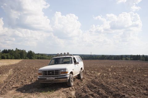 1993 Ford Bronco - A white Ford Bronco is parked in a freshly plowed field under a cloudy sky. The vehicle has four auxiliary lights mounted on its roof rack.