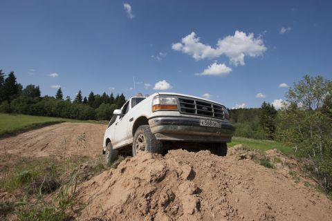 1993 Ford Bronco - A white Ford Bronco is parked on a dirt mound with a dirt road and trees in the background under a blue sky with white clouds.