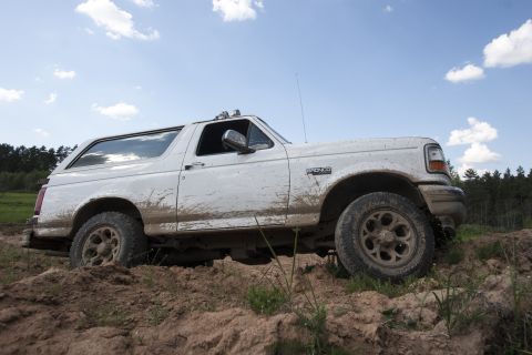 1993 Ford Bronco - A white, mud-splattered 1993 Ford Bronco is shown from a low angle, driving off-road on a dirt path with trees in the background.