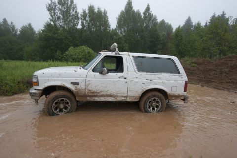 1993 Ford Bronco - A white, mud-splattered 1993 Ford Bronco is stuck in a muddy puddle with trees and a grassy field in the background.