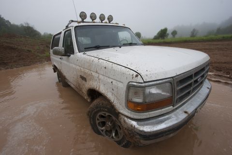 1993 Ford Bronco - A white Ford Bronco is stuck in a muddy puddle, covered in mud. The vehicle has four auxiliary lights mounted on the roof rack.