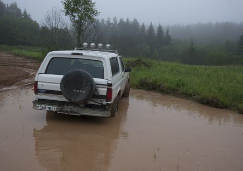 1993 Ford Bronco - A white Ford Bronco is driving through a muddy puddle on a dirt road, with a forest in the background.
