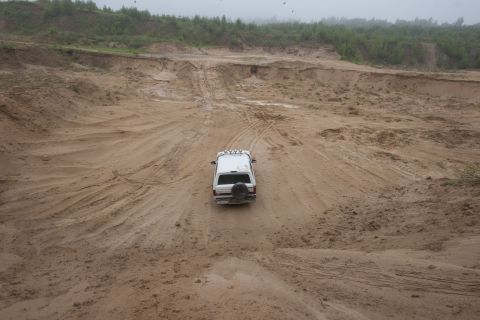1993 Ford Bronco - A white 1993 Ford Bronco is driving on a sandy, rutted track in a quarry. The vehicle has a spare tire mounted on the rear and auxiliary li…
