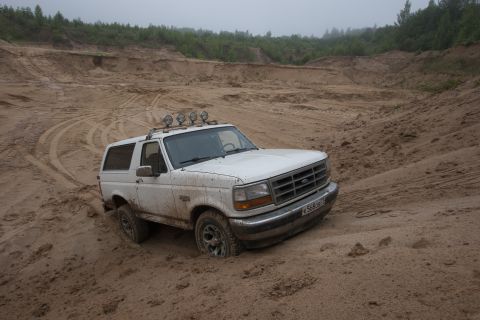 1993 Ford Bronco - A white 1993 Ford Bronco is stuck in a sandy quarry, with its front tires partially buried in the loose sand. The vehicle has a light bar m…