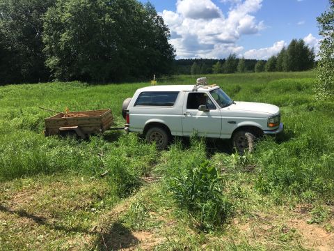 1993 Ford Bronco - A white 1990s Ford Bronco is parked in a grassy field, towing a small utility trailer filled with dirt and tools. The vehicle has auxiliary…