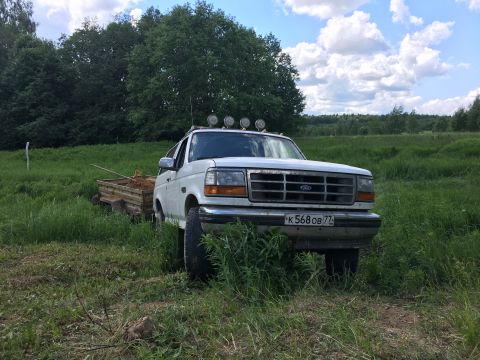 1993 Ford Bronco - A white 1990s Ford Bronco is parked in a grassy field next to a trailer filled with dirt. The truck has four auxiliary lights mounted on th…