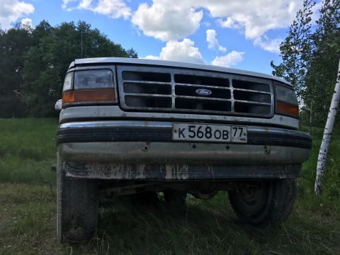 1993 Ford Bronco - The front view of a white Ford Bronco is shown parked outdoors in a grassy area with trees and a cloudy blue sky in the background. The veh…