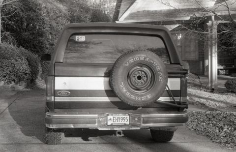 1988 Ford Bronco - Rear view of a dark-colored Ford Bronco with a spare tire mounted on the back. The vehicle is parked on a paved surface next to a house.