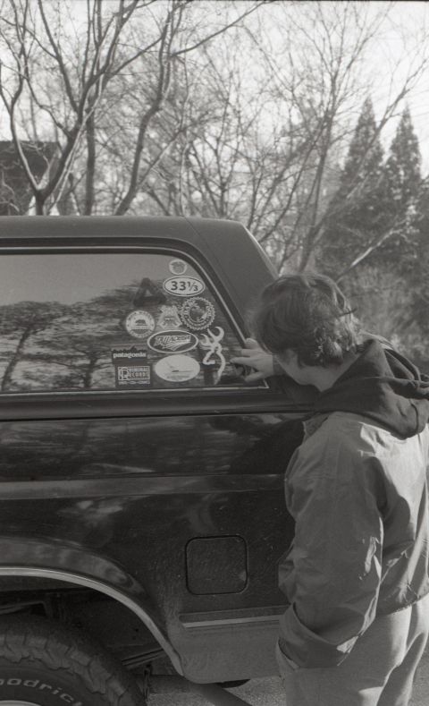 1988 Ford Bronco - A person leans against the rear quarter panel of a dark-colored SUV, looking at stickers on the rear window. The SUV has a camper shell and…