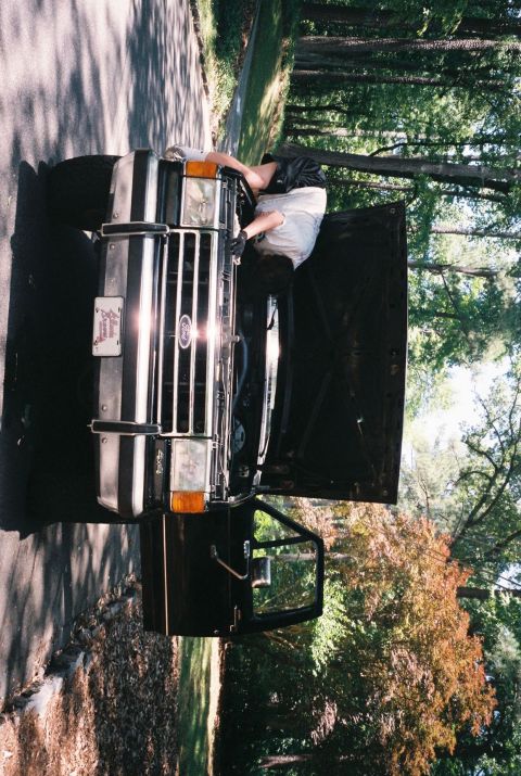 1988 Ford Bronco - A person wearing black shorts and a light-colored t-shirt works on the engine of a black Ford Bronco with its hood open. The vehicle is par…