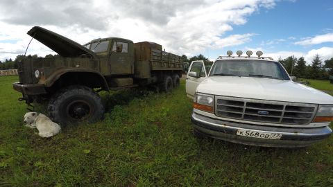 1993 Ford Bronco - A white Ford pickup truck with its driver's side door open is parked next to a large, olive-drab military-style truck with its hood open. A…
