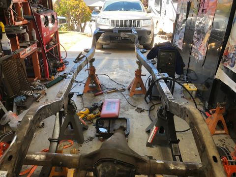 1992 Jeep Wrangler - A vehicle frame is supported by jack stands in a garage setting, with welding equipment and tools nearby. A Jeep Grand Cherokee is visible…