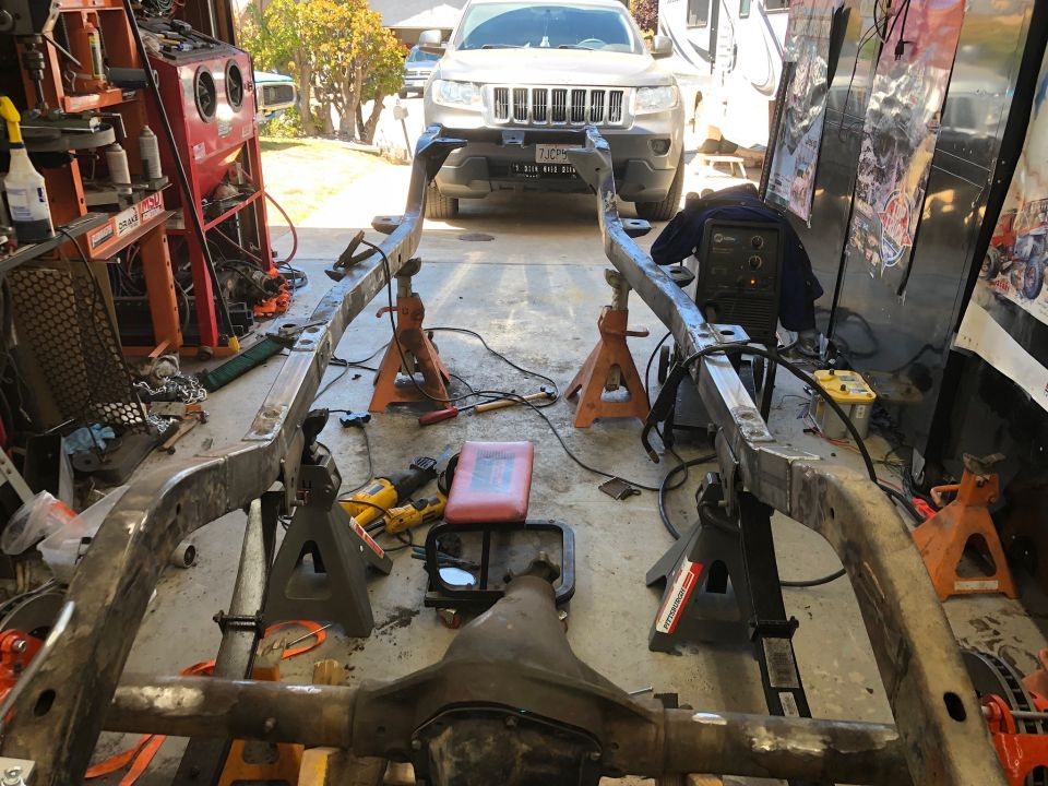 Vehicle frame on jack stands in garage with welding equipment and Jeep Grand Cherokee in background.