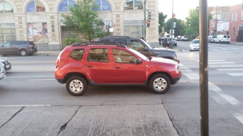 Red Dacia Duster SUV parked on street, with a dark SUV behind it.