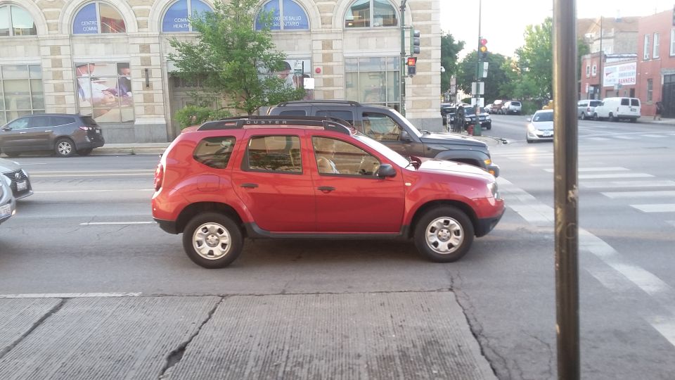 Red Dacia Duster SUV parked on street, with a dark SUV behind it.