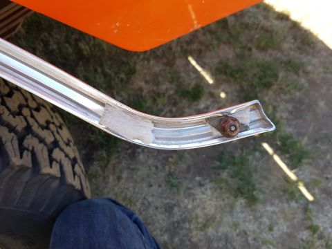 1978 Ford Bronco - A close-up view of a chrome trim piece attached to an orange vehicle body, with a tire and a person's leg in jeans visible. The trim piece…