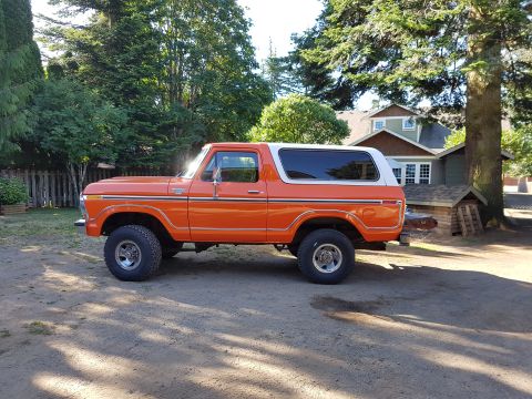 1978 Ford Bronco - A bright orange 1978 Ford Bronco with a white roof is parked on a dirt driveway in front of a house and trees. The classic SUV is shown fro…