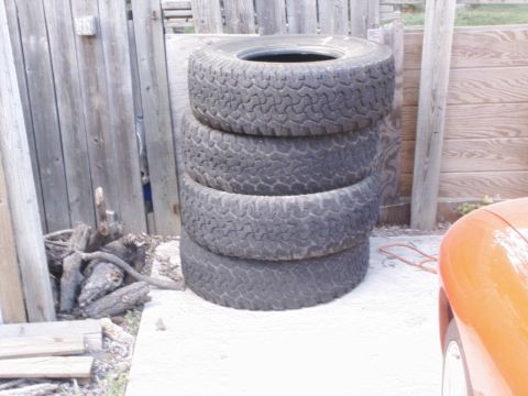 1984 Ford Bronco - A stack of four used off-road tires sits outdoors next to a wooden fence.